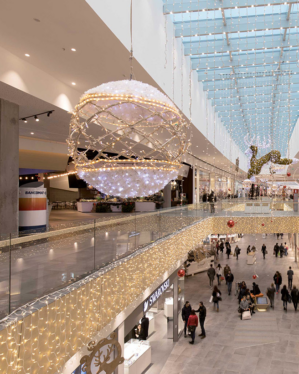 Shoppingcenter med overdådig juleudsmykning, herunder en stor hængende kugleformet dekoration med LED-lys og sneeffekt. Loftet og gelænderet er dekoreret med lyskæder, mens et oplyst rensdyr hænger i baggrunden.