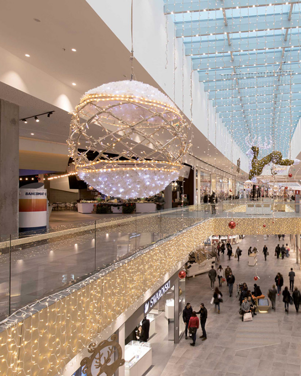 Shoppingcenter med overdådig juleudsmykning, herunder en stor hængende kugleformet dekoration med LED-lys og sneeffekt. Loftet og gelænderet er dekoreret med lyskæder, mens et oplyst rensdyr hænger i baggrunden.