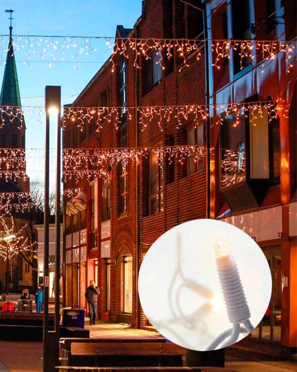 Bygade dekoreret med hængende LED-lysgardiner i varm hvid, skaber en hyggelig atmosfære ved skumring. Nærbillede viser en varm hvid LED-pære på en hvid ledning, anvendt i udsmykningen.