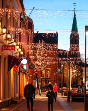Stemningsfuld bygade ved skumring, dekoreret med hængende LED-lysgardiner og oplyste træer i varm hvid. Folk går langs gaden, der er oplyst af juledekorationer, med en kirke i baggrunden.