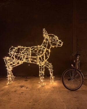 "En lysskulptur af et rådyr opbygget af LED-lys, der udsender en varm glød. Skulpturen står på et betonunderlag i et mørkt rum, og en sort cykel er placeret til højre for skulpturen, hvilket tilføjer en kontrast til scenen."