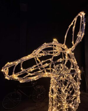 Nærbillede af hovedet på en lysende hunhjortskulptur fremstillet af en gennemsigtig metalramme med varme gule LED-lys. En cykel ses svagt i baggrunden mod en mørk væg.