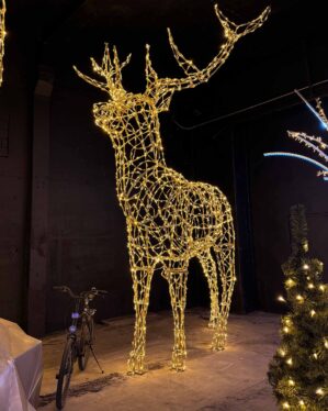 Et majestætisk 3D-krondyr fremstillet i guld-pulverlakeret aluminium og dekoreret med varm hvid LED-lys, der skaber en imponerende og elegant belysning. Den store figur, placeret i et mørkt rum, fremhæver detaljerne i konstruktionen og det strålende lys. En cykel ved siden af giver perspektiv til krondyrets imponerende størrelse, mens dekorationer i baggrunden understreger den festlige atmosfære.