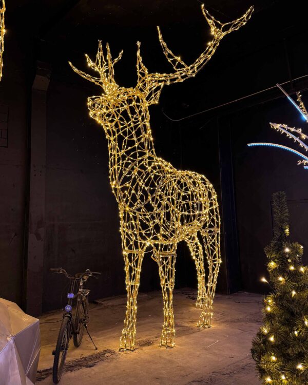 Et majestætisk 3D-krondyr fremstillet i guld-pulverlakeret aluminium og dekoreret med varm hvid LED-lys, der skaber en imponerende og elegant belysning. Den store figur, placeret i et mørkt rum, fremhæver detaljerne i konstruktionen og det strålende lys. En cykel ved siden af giver perspektiv til krondyrets imponerende størrelse, mens dekorationer i baggrunden understreger den festlige atmosfære.