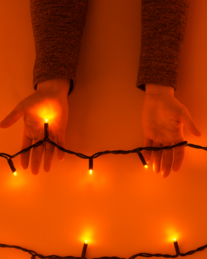 To hænder holder en sort lyskæde med orange LED-lys, som lyser varmt op mod en orange baggrund og skaber en hyggelig og stemningsfuld atmosfære.