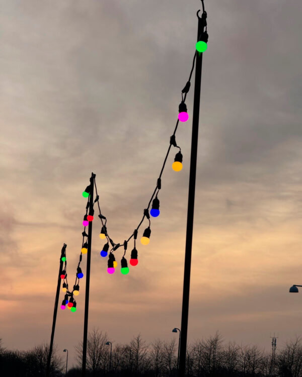 Høje sorte aluminium stativer som holder farverige lyskæder strakt ud i en dekorativ belysning mod en smuk solnedgang. Perfekt til udendørs arrangementer og stemningsfuld aftenbelysning.