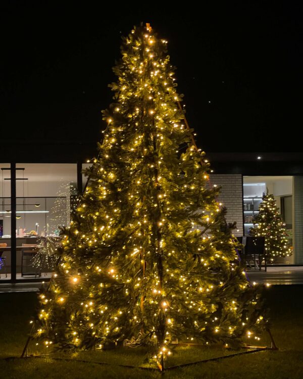 Udendørs juletræ med 2D flader og  med varm hvid belysning står opstillet i have ved moderne hus. Indendørs ses et mindre pyntet træ gennem store glaspartier.