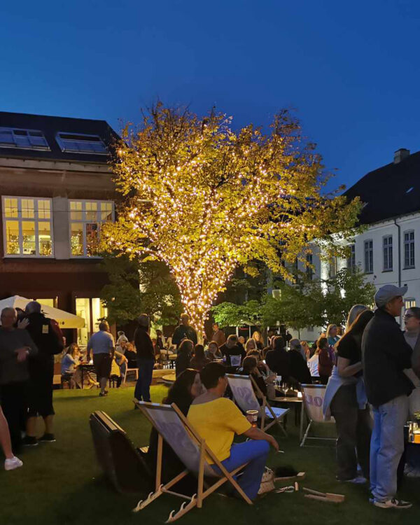 Stort træ med lyskæder i trækronen bag en stor menneskemængde til sommerfest i bymiljø.