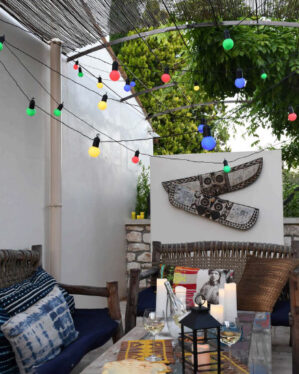 Udendørs loungeområde med farvede festoon-lyskæder i rød, gul, blå og grøn. Skaber hyggelig og dekorativ stemning til sommeraftener, caféer og private terrasser.