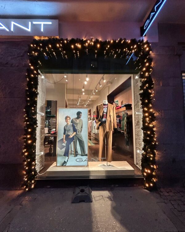 Butiksfacade dekoreret med mørkegrøn gran og varm hvid LED-belysning omkring vinduet. Skaber en elegant og indbydende julestemning foran GANT-butikken i aftentimerne.