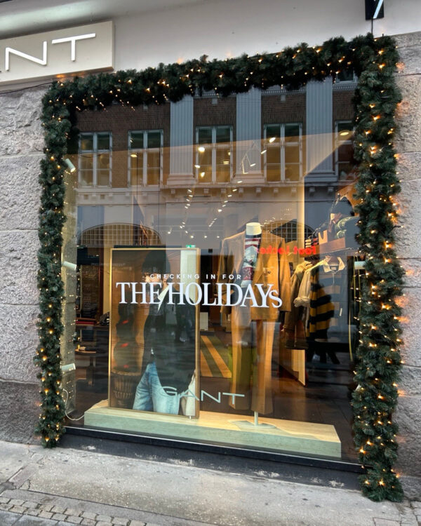 GANT-butik med facade dekoreret i tæt, mørkegrøn gran og varm hvid LED-belysning. Skaber et eksklusivt og festligt udtryk, perfekt til julens butiksvinduer.