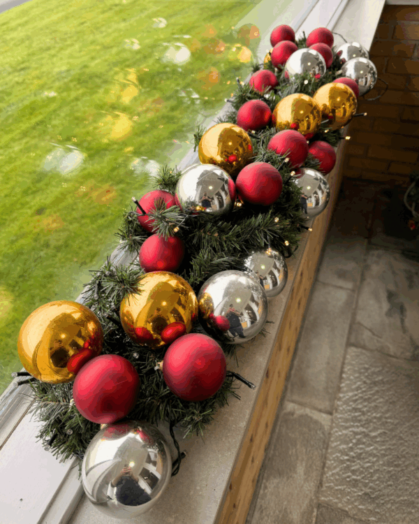 Elegant granguirlande pyntet med røde, guld og sølvfarvede kugler samt varm hvide LED-lys. Perfekt til vindueskarm eller butik for klassisk julestemning.