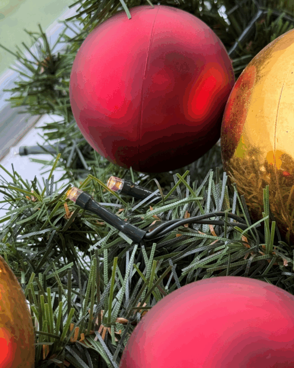 Detaljeret nærbillede af granguirlande med røde og guld julekugler samt varm hvide LED-lys. Viser tekstur, glød og klassisk julestemning i smuk balance.