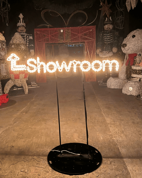 Oplyst "Showroom"-skilt i varm hvid LED på sort platform, placeret indendørs blandt julemotiver, velegnet som stemningsskabende wayfinding og blikfang i professionelle showroom- og butiksområder.