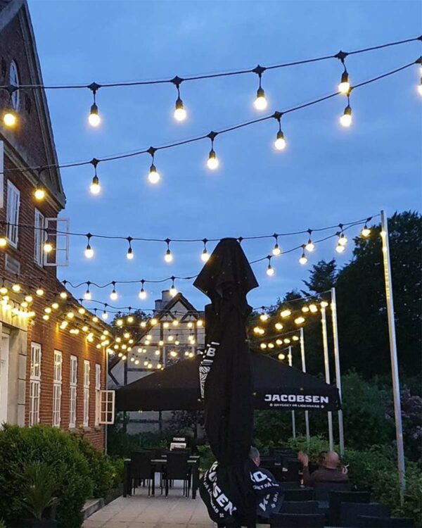 Udendørs terrasse med ophængt party-lyskæde og varme E27 LED-pærer, skaber hyggelig stemning mellem bygninger i aftenlys.