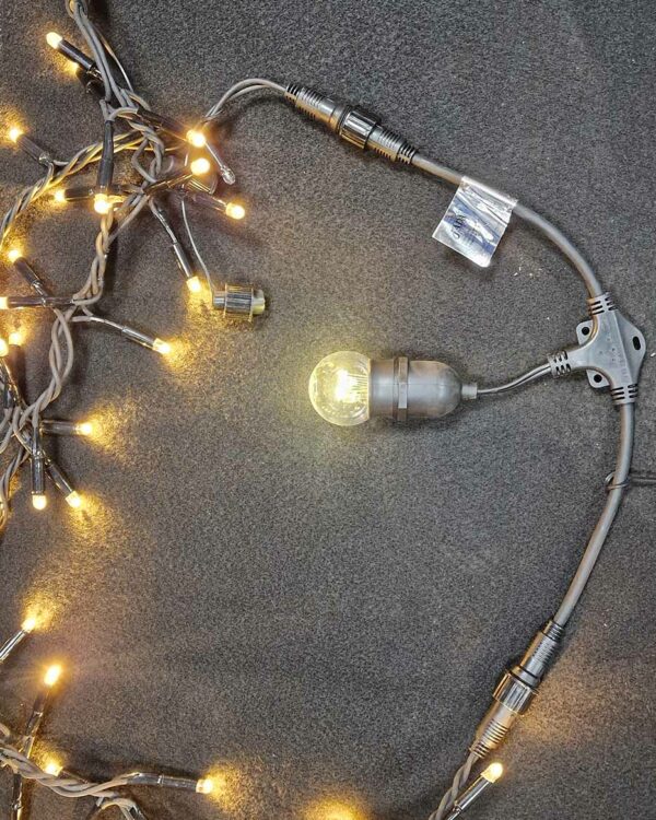Grå T-splitter med E27-fatning til lyskæder, omgivet af varme LED-lys, designet til sikker og fleksibel sammenkobling af udendørs dekorationer.