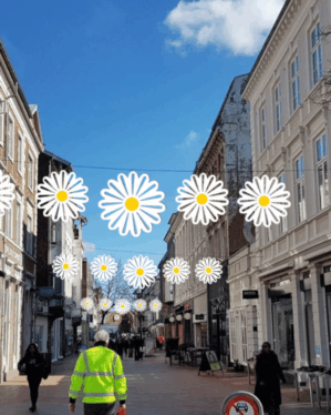 Dekorativ gadeudsmykning med hvide 2D-margueritblomster ophængt over gågade mellem bygninger, skaber sommerlig stemning i byrum med mennesker og butikker.