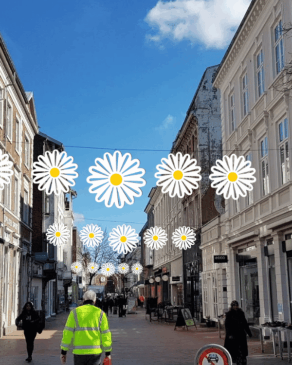 Dekorativ gadeudsmykning med hvide 2D-margueritblomster ophængt over gågade mellem bygninger, skaber sommerlig stemning i byrum med mennesker og butikker.