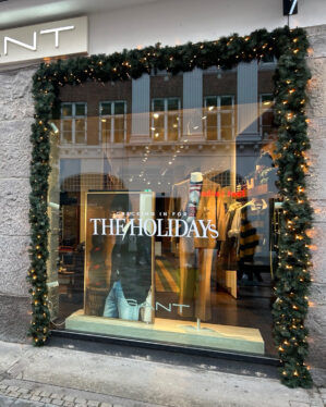 Butiksvindue dekoreret med kunstig granguirlande og varm hvide LED-lys omkring rammen, skaber stemningsfuld juleudsmykning ved indgangen til en tøjbutik i gademiljø.