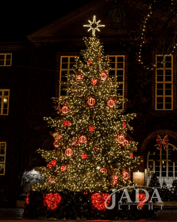Juletræ med tæt varm hvid belysning, røde dekorationer og 2D-topstjerne i varm hvid ropelight, vist foran bygning som stemningsfuld og elegant juleudsmykning.